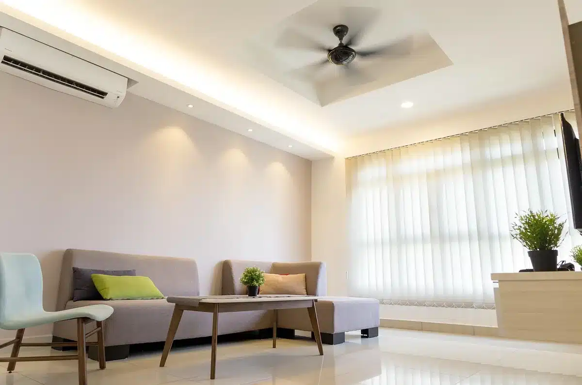 Climatiseur mobile posé près d'une fenêtre dans un salon lumineux, avec une main ajustant la télécommande